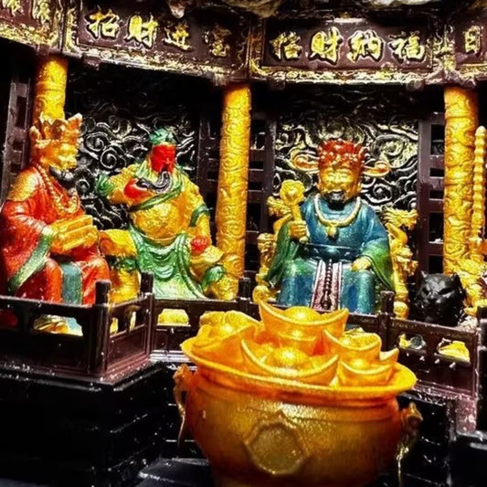 Close-up of three Five Roads God of Wealth deity figurines seated in a miniature shrine diorama, with gold ingot cauldron in foreground and ornate carved columns and blessing banners behind
