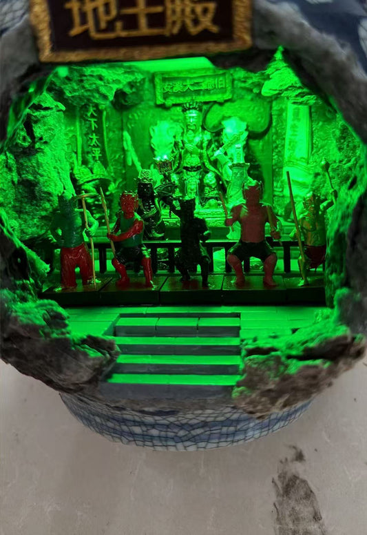 Interior view of a miniature temple diorama through a cave rock opening, featuring five Wu Zu Jiang Jun (五卒将军) figurines seated in a row under vivid green lighting, with a large Tua Di Gong and Tua Ji Ya Pek (大二爷伯) behind. Chinese inscription plaque reading 地主殿 above the entrance
