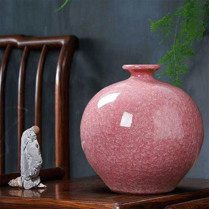 Lifestyle shot of a round pink crackle-glaze pomegranate porcelain vase displayed on a dark wooden furniture surface, with a small grey monk figurine bowing beside it and green foliage visible in the background, shown as a blank custom vase base before shrine cutout modification