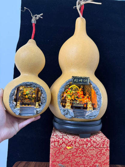 Two natural gourd vessels side by side against a black background — a small gourd held in hand on the left with an illuminated shrine diorama inside, and a larger gourd on the right with a cave cutout revealing an illuminated 财神殿 miniature shrine diorama with deity figurines, displayed on a black round base atop a red brocade gift box
