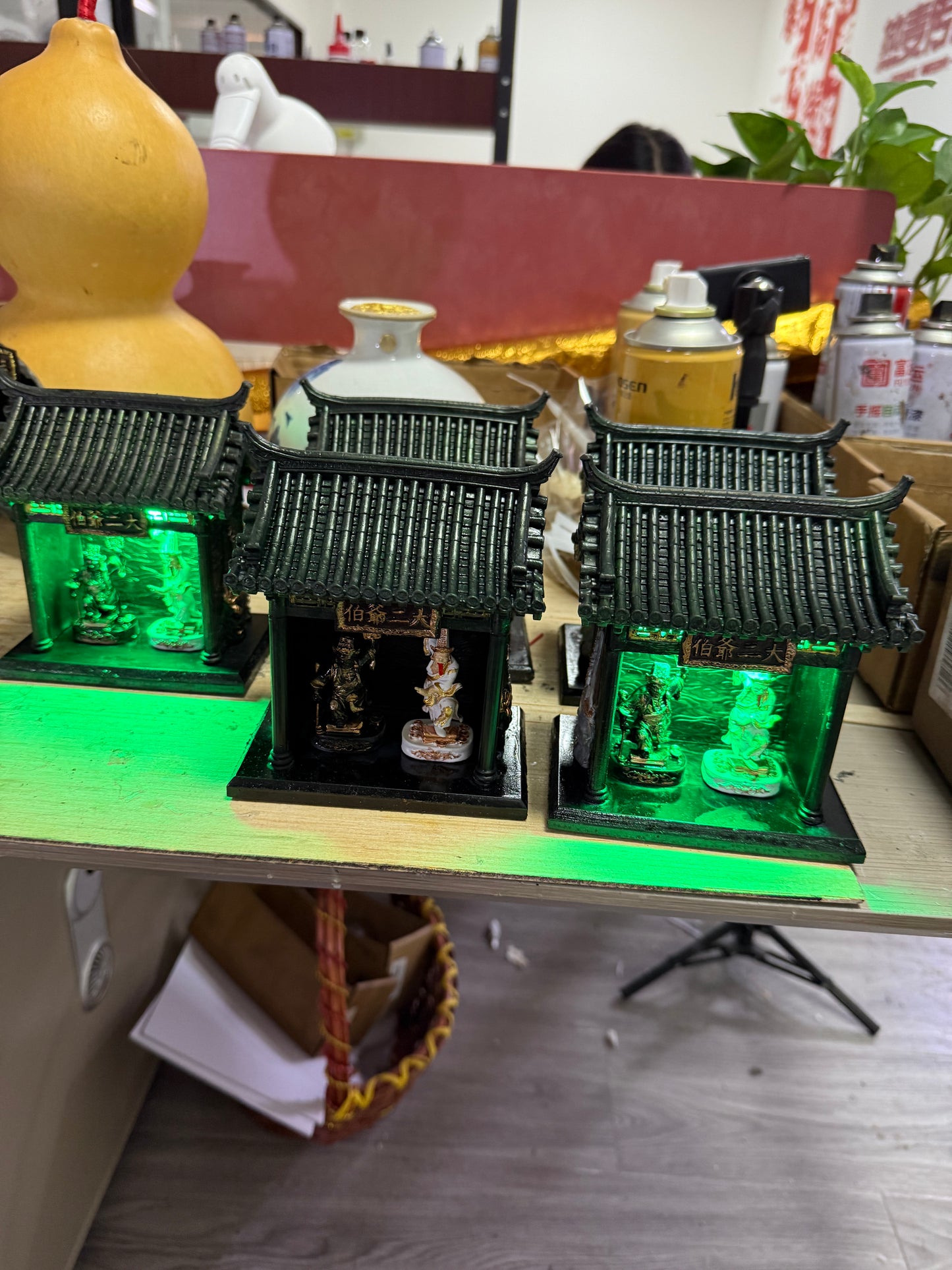 Shrine temple models with Tua Di Ya Pek figurines under green lighting displayed in a workshop setting 
