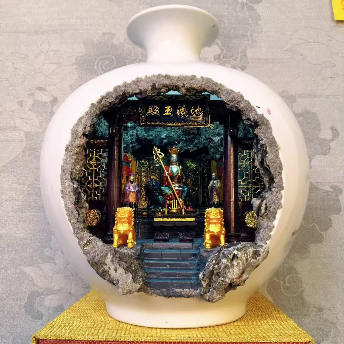 Full front view of a large round white glazed porcelain vase with a wide circular cave cutout revealing an illuminated 地藏王殿 miniature shrine diorama featuring Ksitigarbha Bodhisattva seated on a throne holding a golden ringed staff, flanked by two attendant figurines and two gold Kirin guardians on the altar steps, displayed on a yellow woven mat