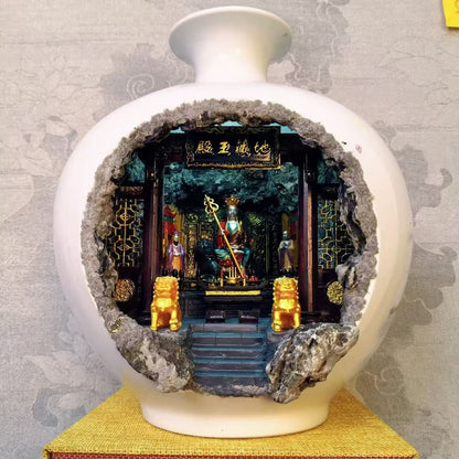 Full front view of a large round white glazed porcelain vase with a wide circular cave cutout revealing an illuminated 地藏王殿 miniature shrine diorama featuring Ksitigarbha Bodhisattva seated on a throne holding a golden ringed staff, flanked by two attendant figurines and two gold Kirin guardians on the altar steps, displayed on a yellow woven mat