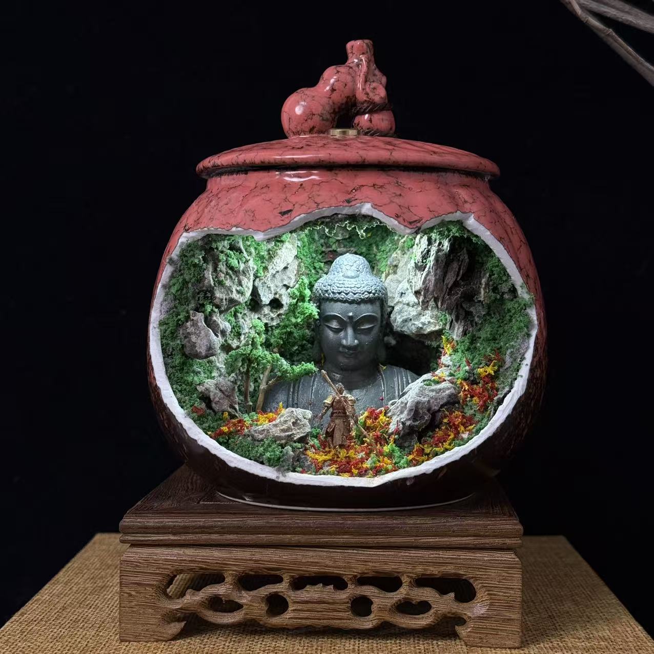 Front view of a round red-pink crackle-glaze lidded ceramic jar, featuring a large oval cave cutout revealing an illuminated miniature diorama of a giant stone Buddha head emerging from a mossy rock cave, with a small armoured Sun Wukong figurine and red and yellow wildflowers at the base, displayed on a carved wooden stand on a burlap mat