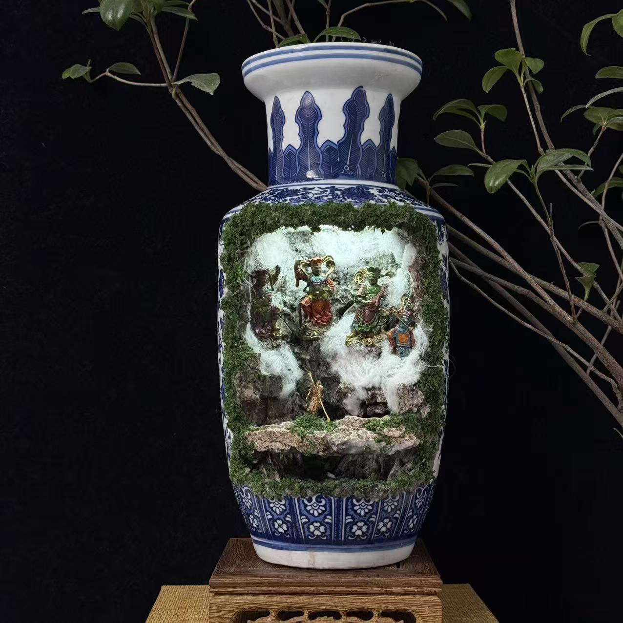 Front view of a tall blue and white porcelain vase with a moss-covered cave cutout revealing an illuminated miniature diorama featuring multiple colourful deity figurines in a battle scene with a golden Sun Wukong figurine at the front, displayed on a carved wooden stand against a black background with plant branches