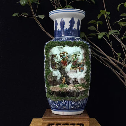 Front view of a tall blue and white porcelain vase with a moss-covered cave cutout revealing an illuminated miniature diorama featuring multiple colourful deity figurines in a battle scene with a golden Sun Wukong figurine at the front, displayed on a carved wooden stand against a black background with plant branches