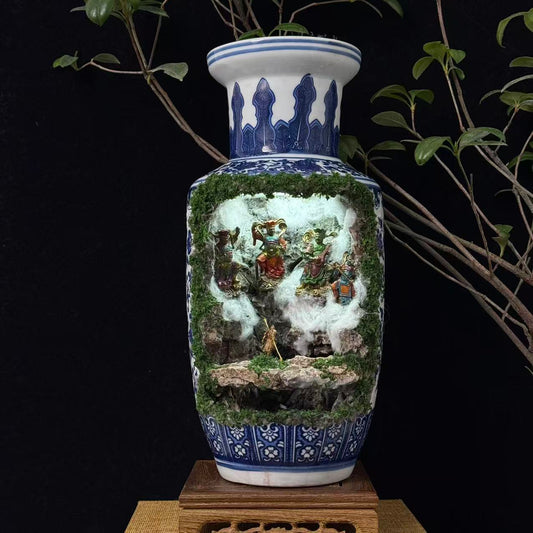 Front view of a tall blue and white porcelain vase with a moss-covered cave cutout revealing an illuminated miniature diorama featuring multiple colourful deity figurines in a battle scene with a golden Sun Wukong figurine at the front, displayed on a carved wooden stand against a black background with plant branches