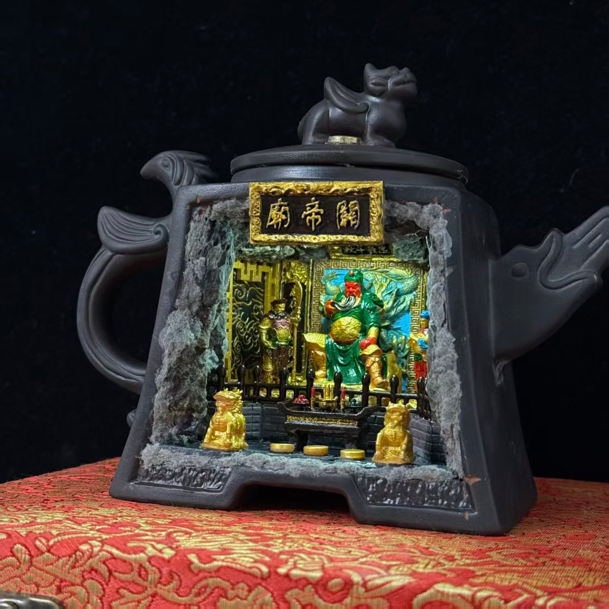 Front view of a dark Yixing-style square ceramic teapot with a Pixiu lid finial and fish-shaped spout and handle, featuring a rectangular cave cutout and a gold 关帝庙 plaque above, revealing an illuminated miniature shrine diorama of Guan Yu seated on a throne in green armour with a red face, flanked by attendant figurines and two gold Pixiu guardians on the altar steps, displayed on a red brocade surface
