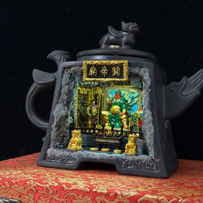 Front view of a dark Yixing-style square ceramic teapot with a Pixiu lid finial and fish-shaped spout and handle, featuring a rectangular cave cutout and a gold 关帝庙 plaque above, revealing an illuminated miniature shrine diorama of Guan Yu seated on a throne in green armour with a red face, flanked by attendant figurines and two gold Pixiu guardians on the altar steps, displayed on a red brocade surface