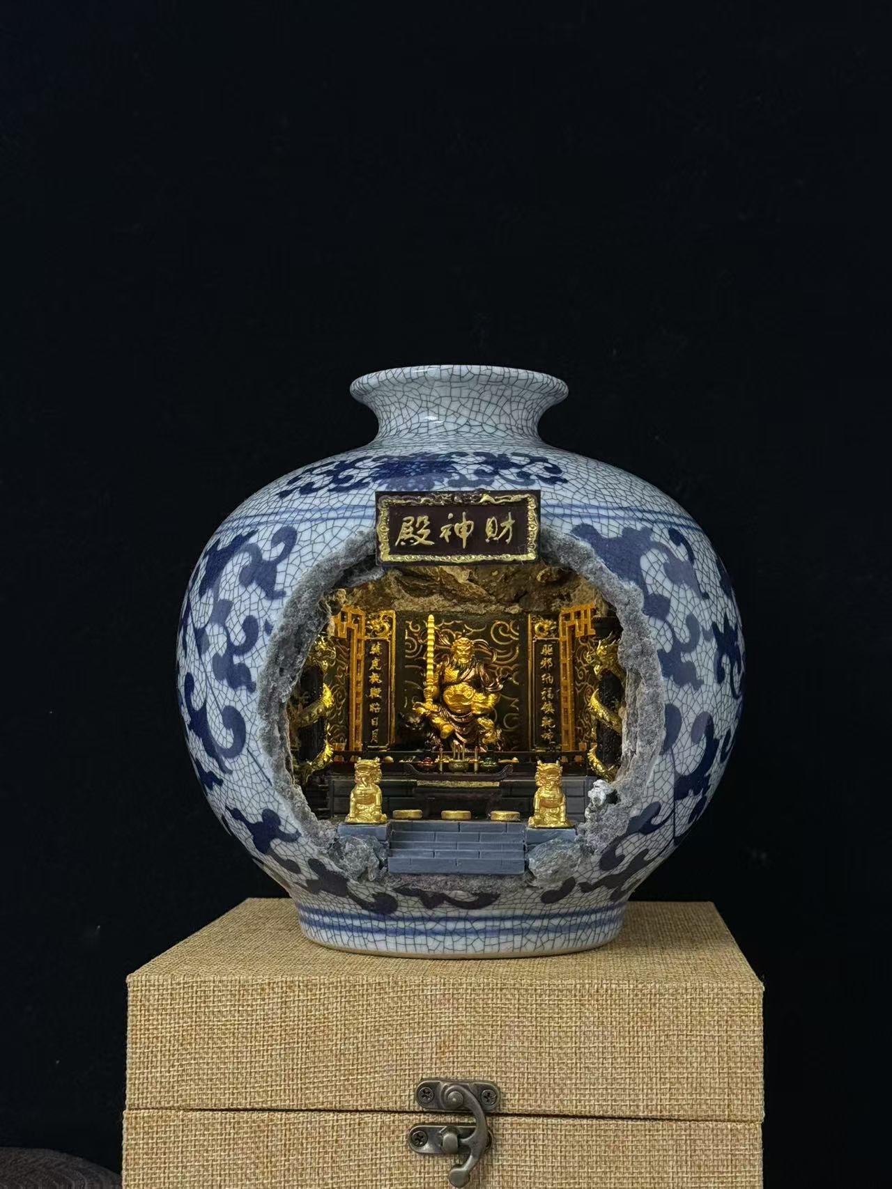 A blue and white porcelain vase with a carved open center revealing a Wu Caishen miniature golden temple scene. The vase is on a beige fabric-covered box against a black background.