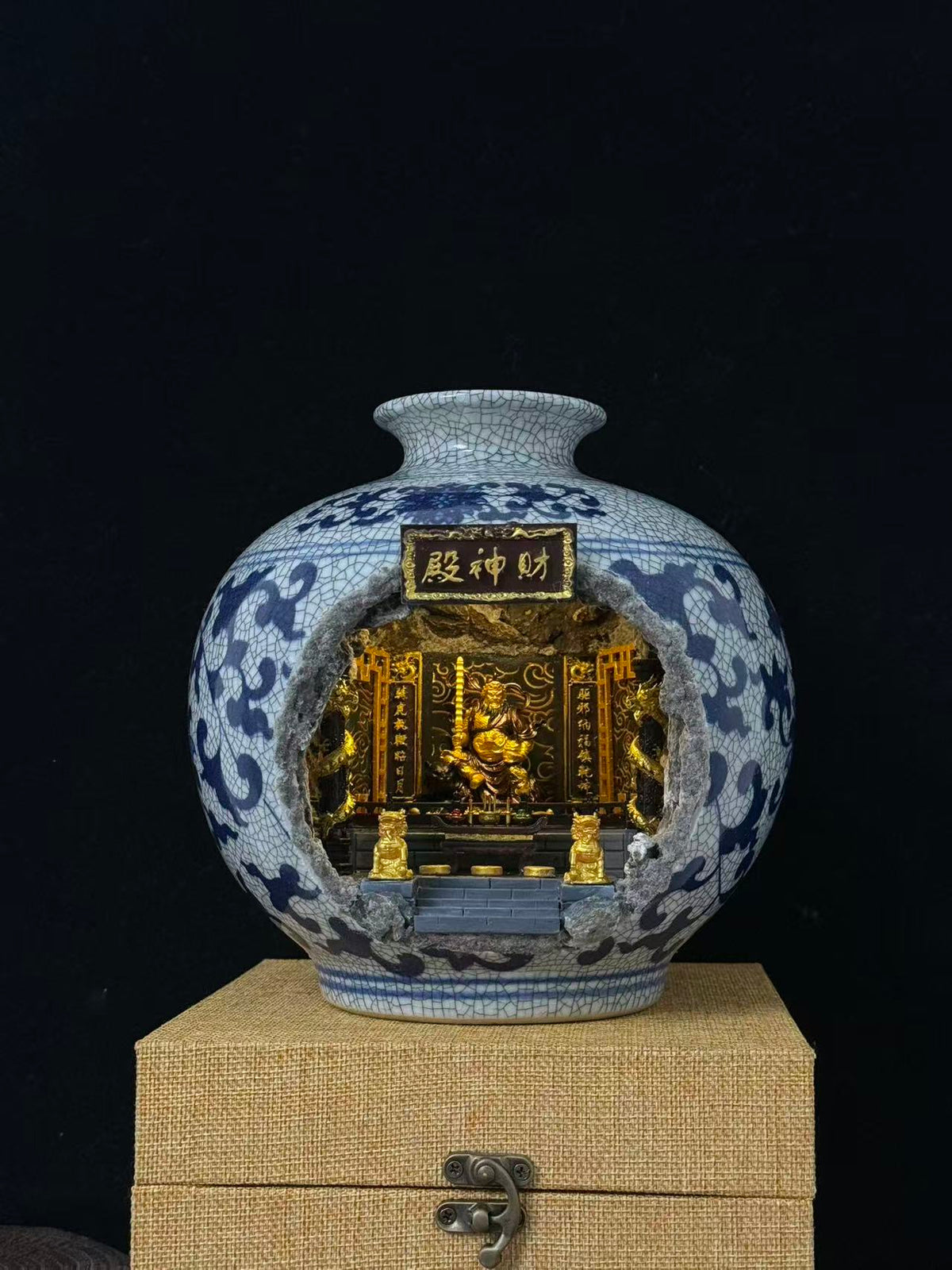 A blue and white porcelain vase with a carved open center revealing a Wu Caishen miniature golden temple scene. The vase is on a beige fabric-covered box against a black background.