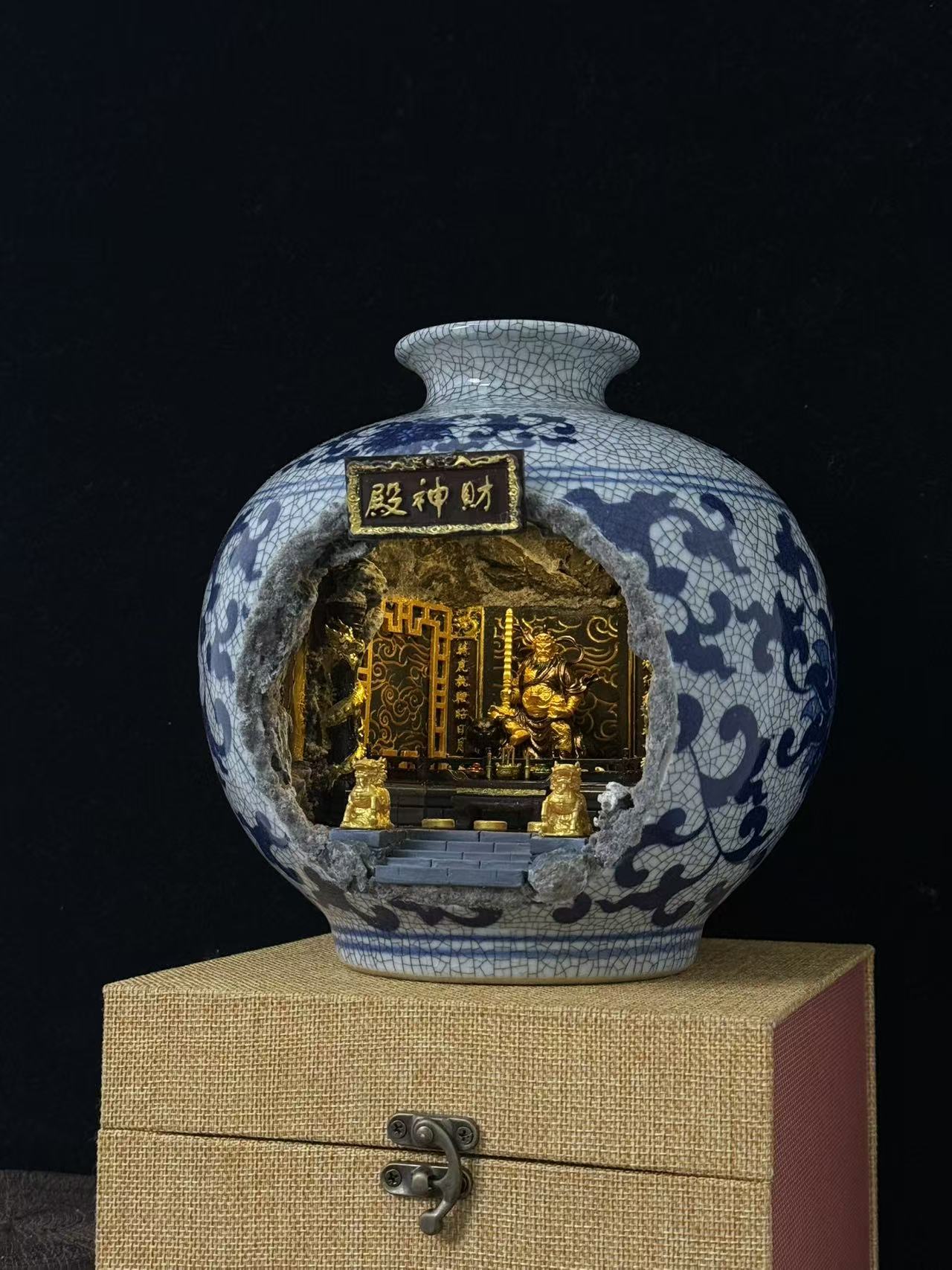 A decorative ceramic vase with a blue-and-white cracked glaze floral pattern and a cutout front displaying a miniature Wu Caishen golden temple scene, displayed on a beige box against a black background.