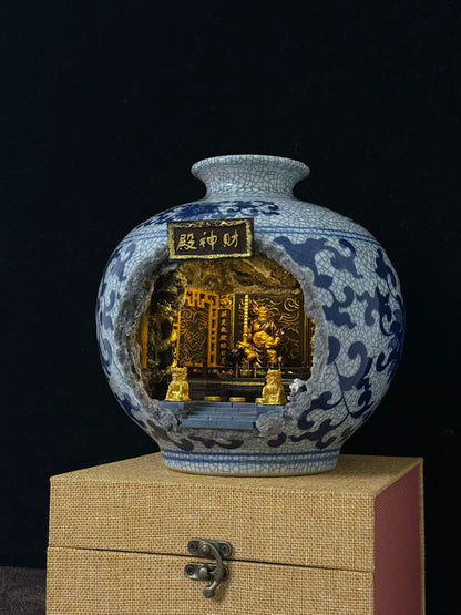 A decorative ceramic vase with a blue-and-white cracked glaze floral pattern and a cutout front displaying a miniature Wu Caishen golden temple scene, displayed on a beige box against a black background.