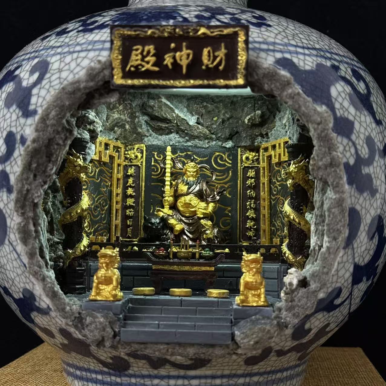 A close-up of a vase with a large broken-front opening, revealing a Wu Caishen miniature golden temple scene inside. The vase is white with blue crackled glaze patterning, and the interior features gold figurines, black panels, dragon motifs, and a central plaque with Chinese characters.