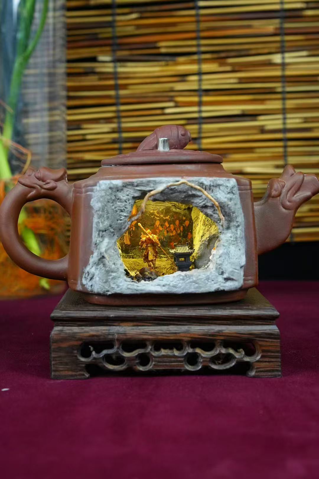 Front view of a brown Yixing-style square ceramic teapot with a cave cutout revealing an illuminated miniature diorama of a Sun Wukong figurine in golden armour standing in an outdoor scene, displayed on a carved wooden stand with a bamboo blind backdrop
