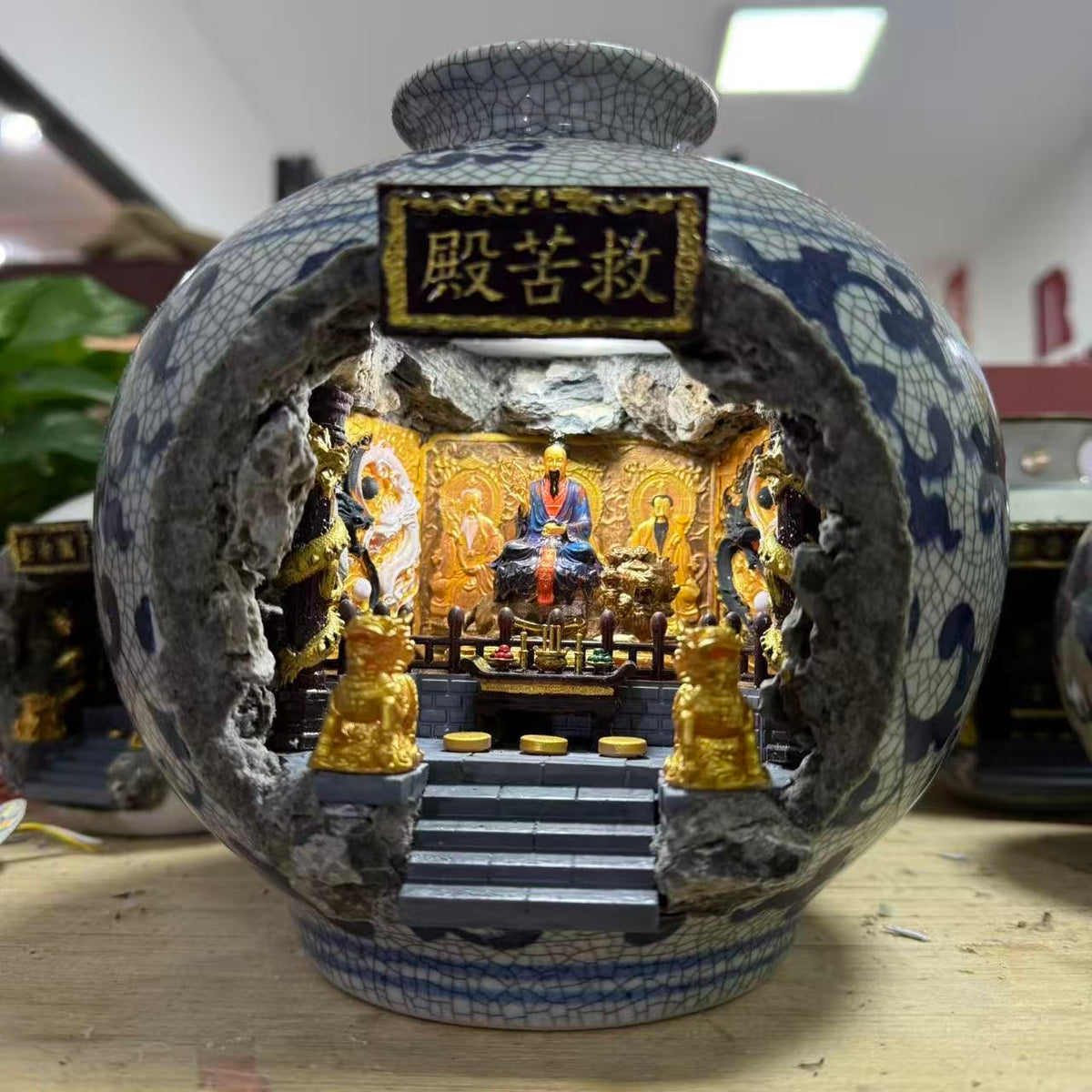 Front view of a dark blue crackle-glaze porcelain vase with a cave cutout revealing an illuminated 救苦殿 miniature shrine diorama featuring Tai Yi Zhen Ren seated on a lotus throne with other Zhen Ten figures on golden carved panels, and two gold Kirin guardians flanking the altar steps, displayed on a wooden workbench