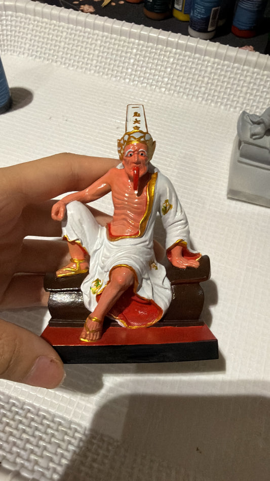 A small painted figurine of a seated tua ya pek figure with peach skin, white robes, and gold details, resting on a red-and-black base. A human hand is holding the left side of the figurine, and the background is a white textured surface with other objects partially visible.