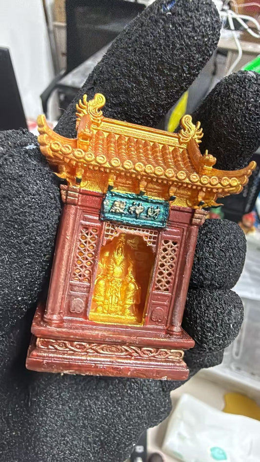 Gloved hand holding a miniature red and gold Chinese temple shrine model with a tiled pagoda roof, lattice window panels, and a teal plaque reading 财神殿, with a gold deity figurine visible inside the shrine opening