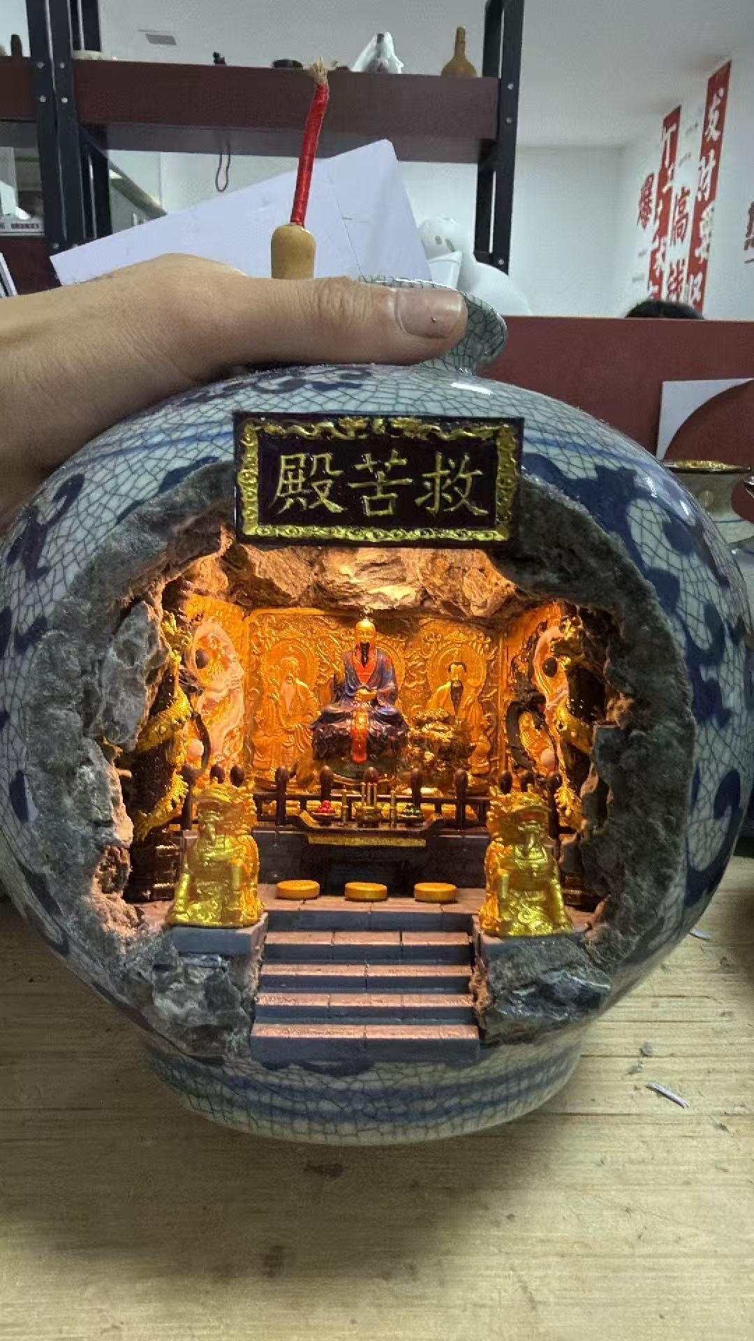 Hand holding a dark blue crackle-glaze porcelain vase showing the illuminated 救苦殿 miniature shrine diorama interior with Tai Yi Zhen Ren seated on a throne under warm orange lighting, with other Zhen Ren figures on golden carved panels and two gold Kirin guardians flanking the altar steps