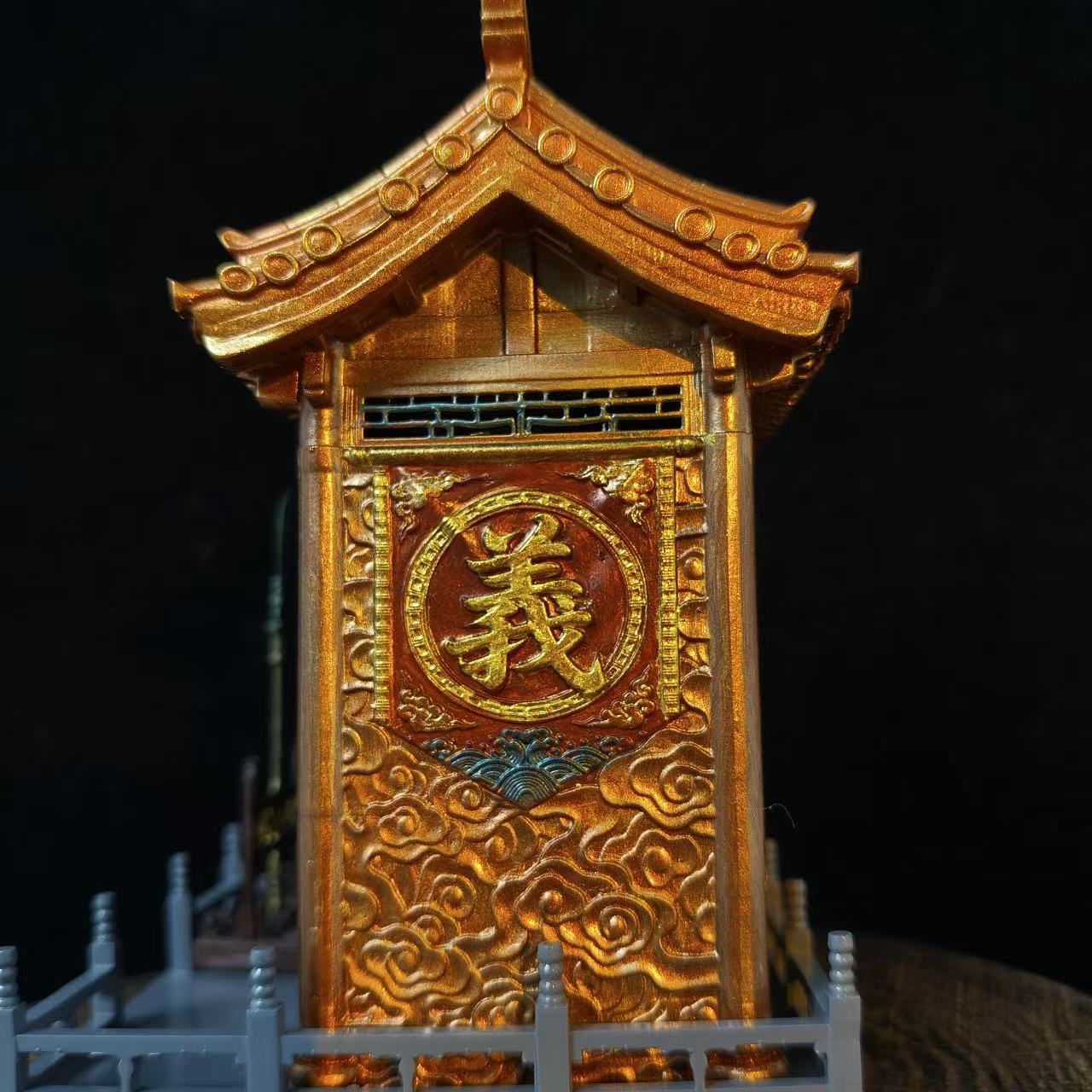 Close-up of the right side panel of a custom miniature car shrine model against a black background, featuring a gold embossed 义 character in a circular frame on a red panel with gold cloud carvings below and pagoda roof corner visible above