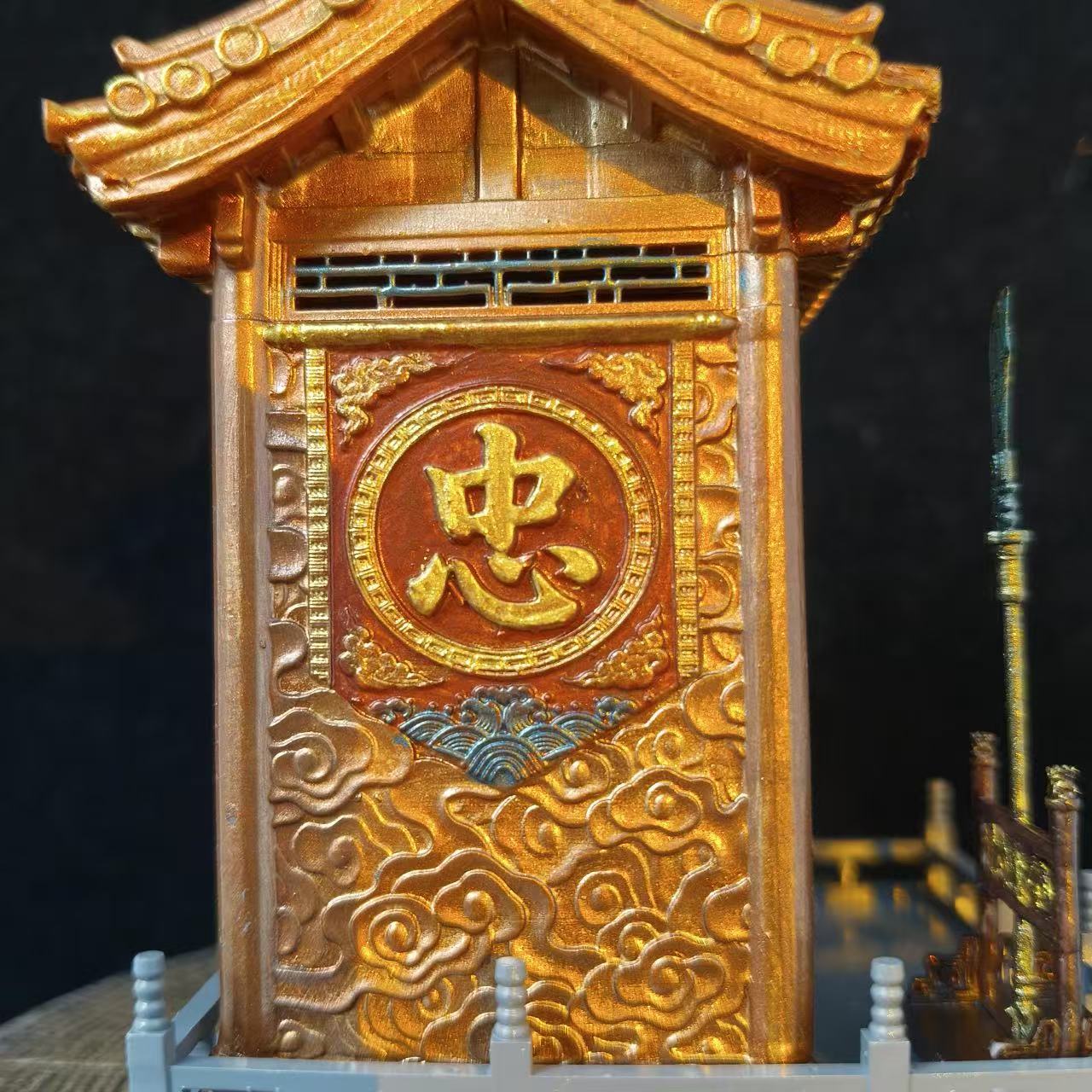 Close-up of the left side panel of a custom miniature car shrine model against a black background, featuring a gold embossed 忠 character in a circular frame on a red panel with gold cloud carvings, with a weapon spear visible at the right edge