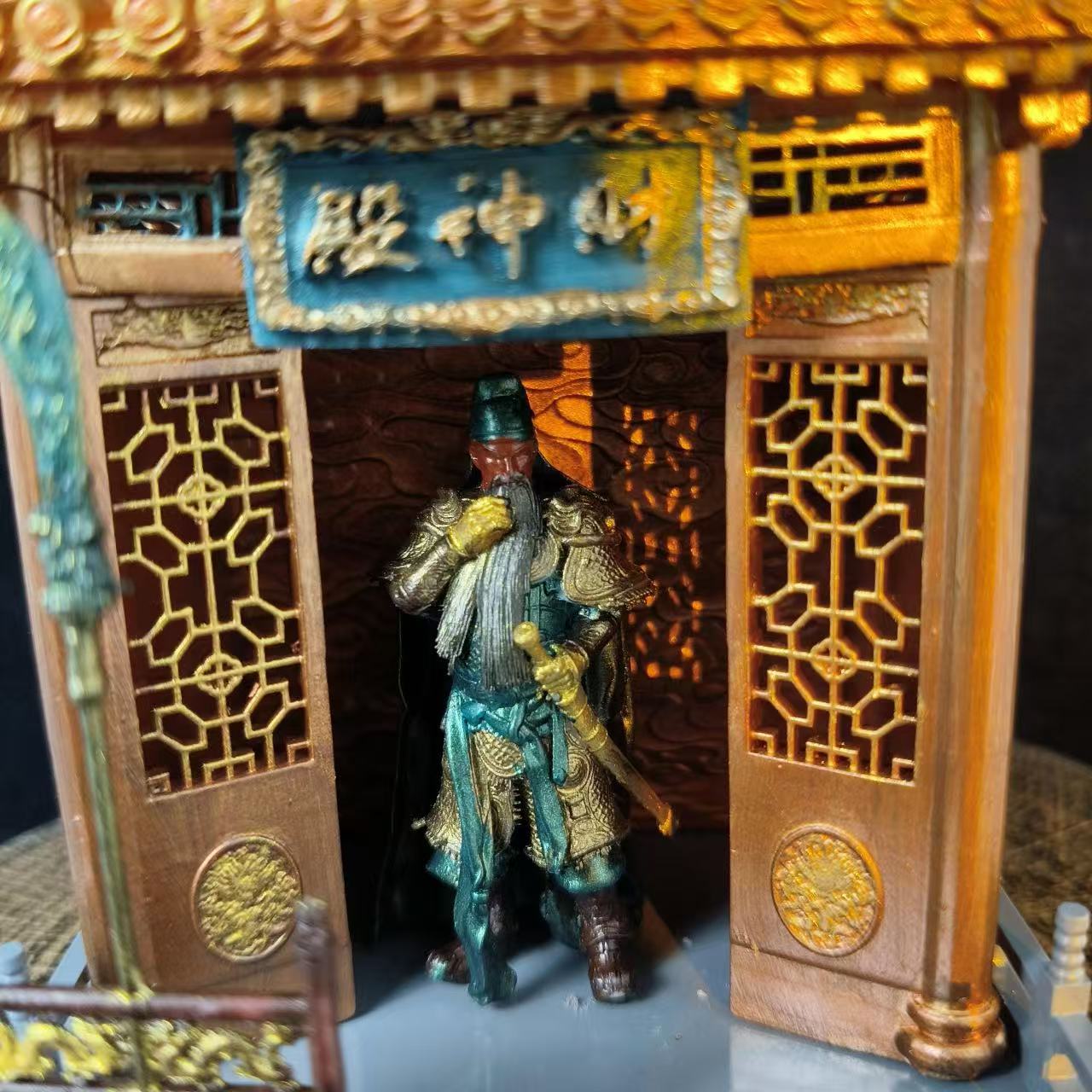 Close-up front view of a custom miniature shrine model featuring a Guan Yu figurine in golden-green armour standing in the doorway, with gold lattice side panels and a teal plaque reading 财神殿 above the entrance