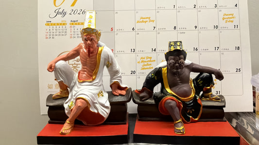 Two Tua Di Ya Pek (大二伯伯) deity figurines displayed side by side — white-robed figurine on the left and dark-robed figurine on the right, photographed in front of a July 2026 calendar