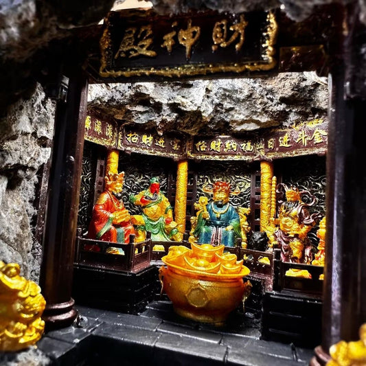 Interior view of a miniature shrine hall diorama featuring the Five Roads God of Wealth seated around a gold ingot cauldron, with carved dragon-cloud wall panels and Chinese blessing inscriptions above