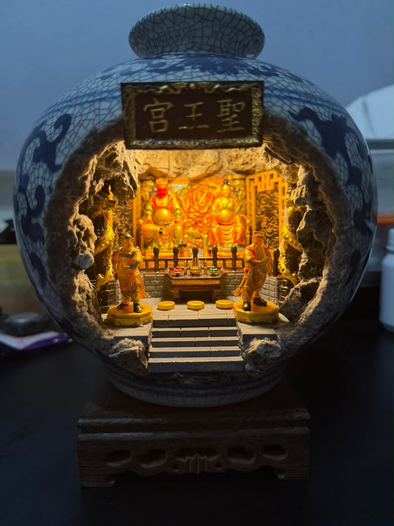 Front view of a blue and white crackle-glaze porcelain vase with a cave cutout revealing an illuminated 圣王宫 miniature shrine diorama, showing Guangze Zunwang and Miao Yin Xian Zi seated on thrones with two gold attendant figurines flanking the altar steps, displayed on a carved wooden stand against a dark background