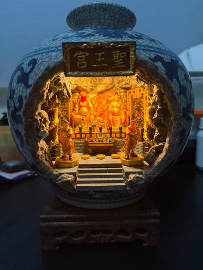 Front view of a blue and white crackle-glaze porcelain vase with a cave cutout revealing an illuminated 圣王宫 miniature shrine diorama, showing Guangze Zunwang and Miao Yin Xian Zi seated on thrones with two gold attendant figurines flanking the altar steps, displayed on a carved wooden stand against a dark background