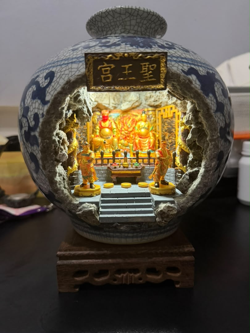 Blue and white crackle-glaze porcelain vase with 圣王宫 miniature shrine diorama inside photographed under natural cooler lighting on a dark desk, showing the full exterior vase form and cave opening with two flanking attendant figurines
