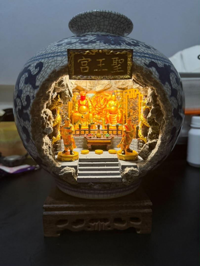 Blue and white crackle-glaze porcelain vase with illuminated 圣王宫 miniature shrine diorama inside, displayed on a carved wooden stand on a dark desk, showing the full vase form with cave opening and two flanking attendant figurines