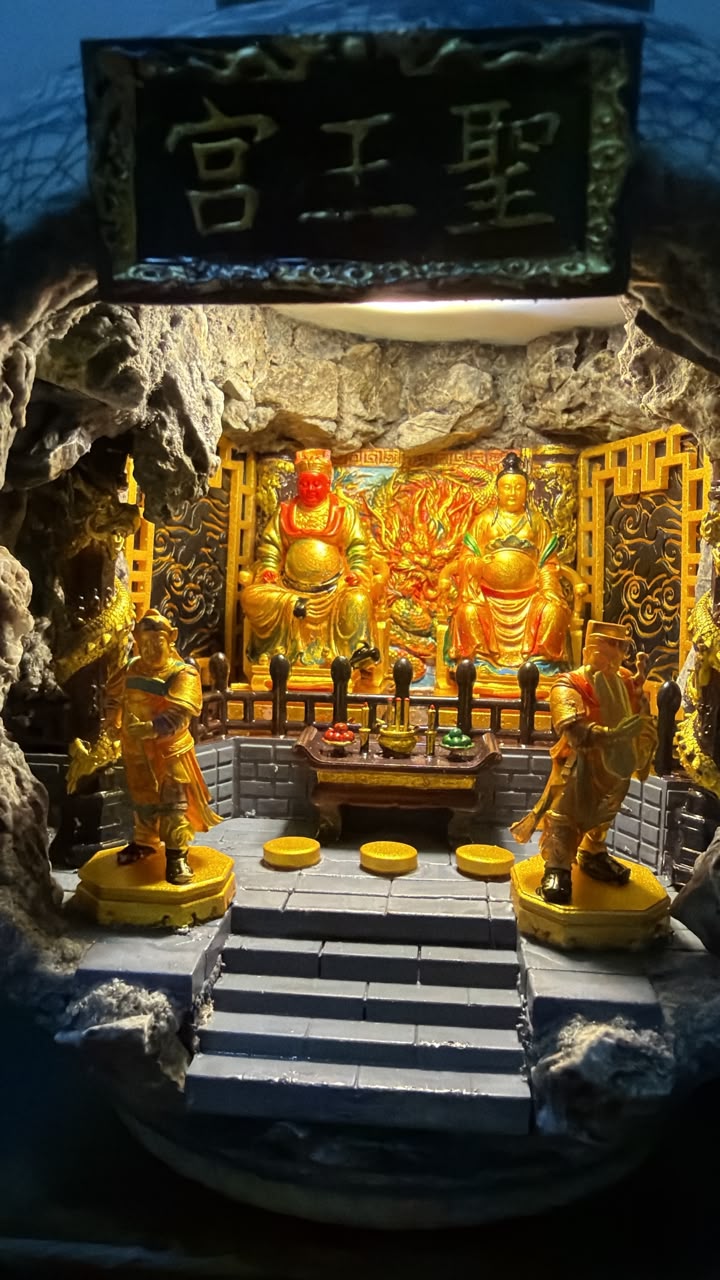Wide-angle interior view of the 圣王宫 miniature shrine diorama under cool blue ambient lighting, showing Guangze Zunwang and Miao Yin Xian Zi on their thrones with two flanking attendant figurines and cave rock arch framing