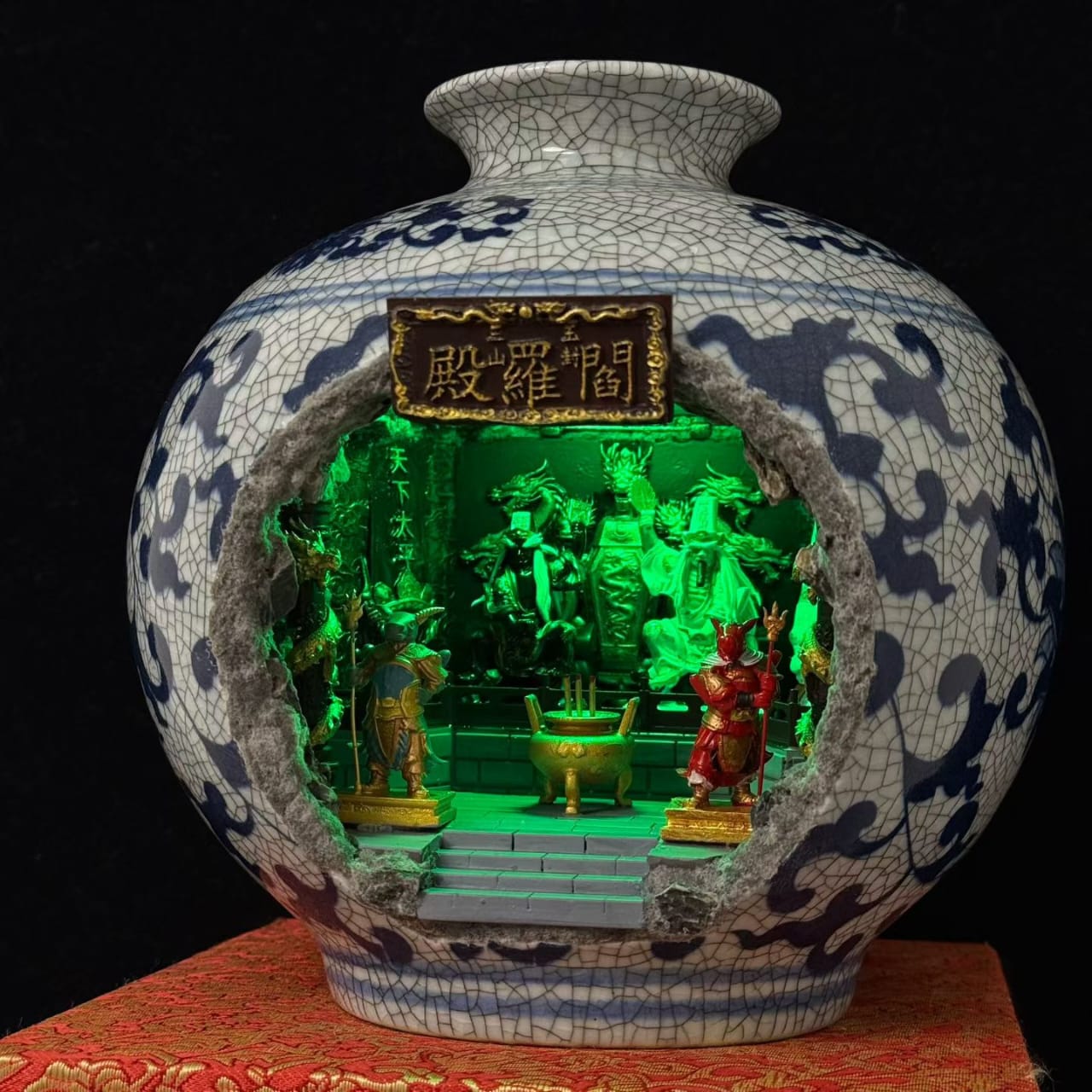 A blue and white porcelain vase with a cutout front revealing a miniature illuminated shrine diorama featuring Tua Di Ya Pek, displayed on a burlap mat against a black background