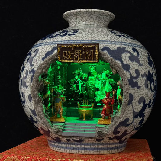 A blue and white porcelain vase with a cutout front revealing a miniature illuminated shrine diorama featuring Tua Di Ya Pek, displayed on a burlap mat against a black background