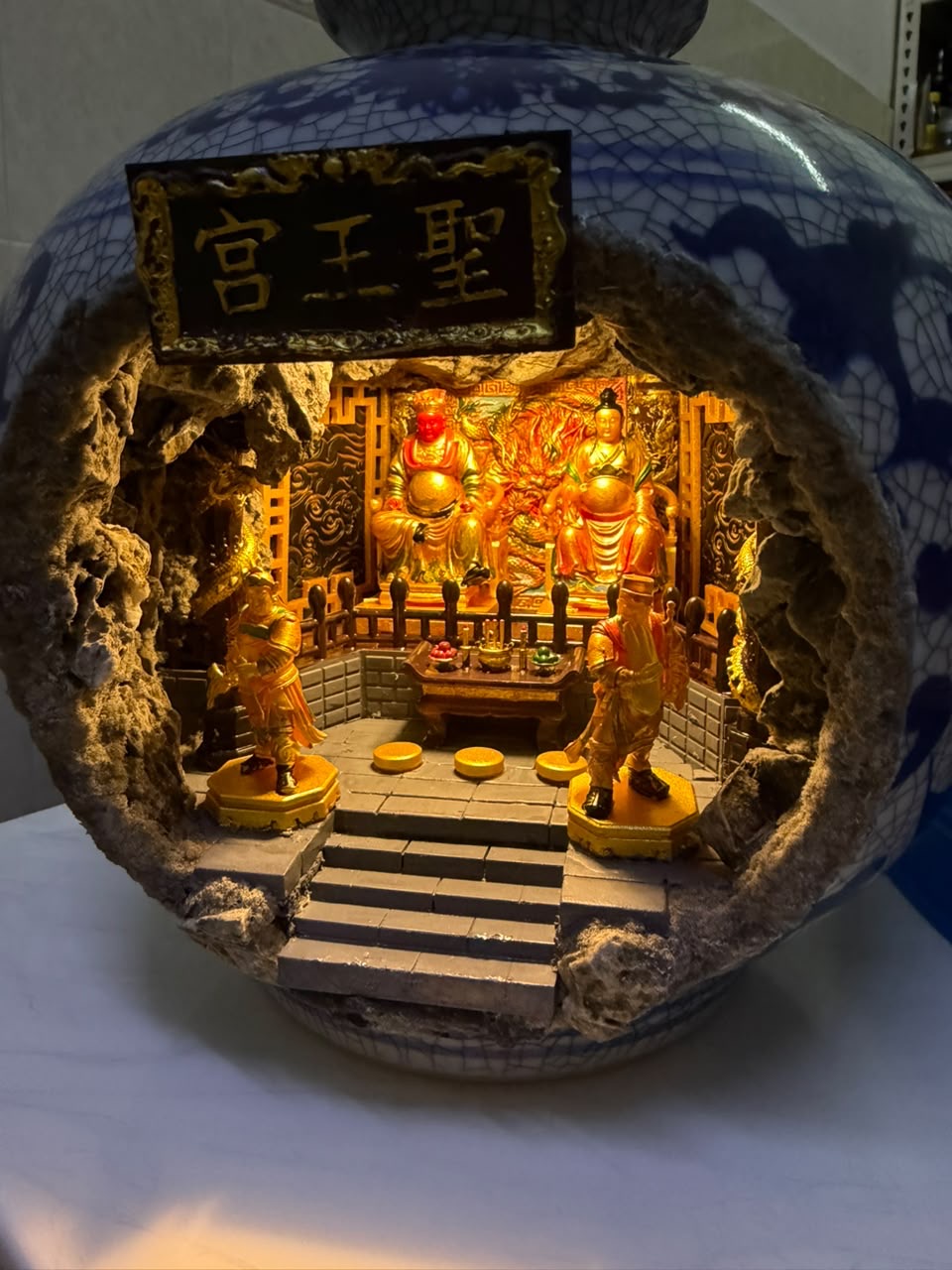 Front view of a blue and white crackle-glaze porcelain vase with a cave cutout revealing an illuminated 圣王宫 miniature shrine diorama, displayed on a white surface with two attendant figurines flanking the altar steps