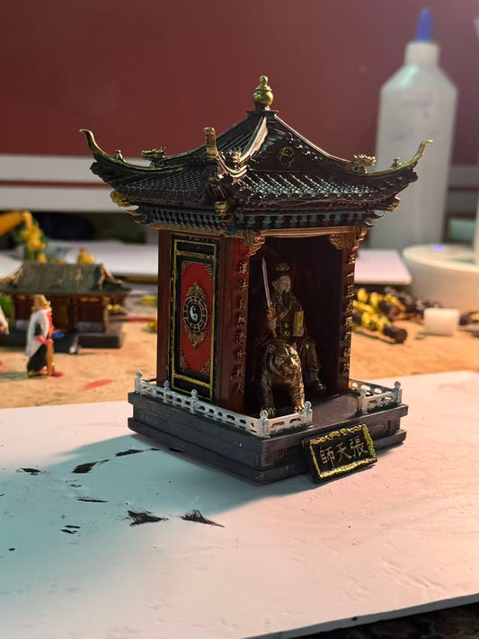 Full view of a custom miniature shrine model with a dark green pagoda roof and gold finial, featuring a Zhang Tianshi deity figurine with a tiger inside the shrine, a red Yin-Yang panel on the left wall, and a name plaque reading 张天师 at the base, photographed on a workshop table